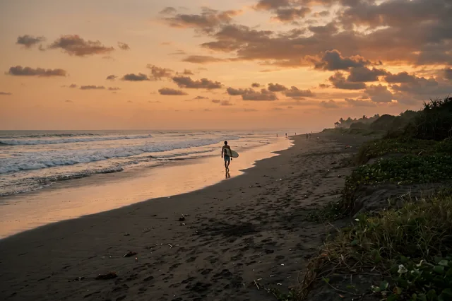 A surfer carrying a board along the dark-sand beach at Pererenan at sunset